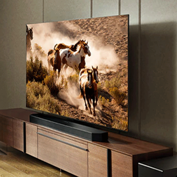 Samsung OLED flat-screen television mounted on a wall above a wooden entertainment console. The television screen shows an image of three horses galloping with a cloud of dust behind them.